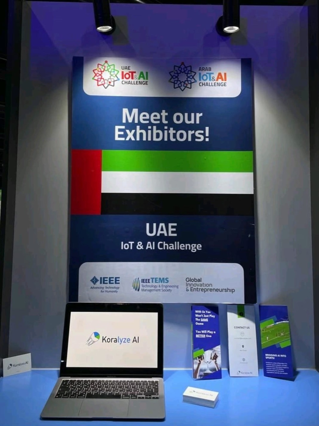 The Koralyze AI booth at the UAE IoT & AI Challenge exhibitor stand at GITEX: a laptop showing the Koralyze logo, brochures and business cards under the event poster.